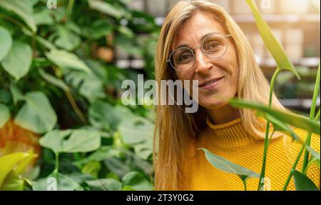 Lächelnde Frau mit Brille inmitten grüner Blumen, die in einem Gartencenter in die Kamera schaut. Einkaufen im Gewächshauskonzept Stockfoto