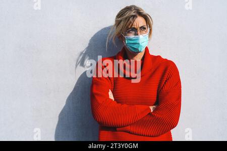 Frau in einem roten Pullover steht vor einem neutralen Hintergrund, trägt eine türkisfarbene Gesichtsmaske und eine runde Brille, mit überkreuzten Armen, weil sie anno ist Stockfoto