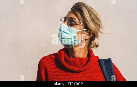 Frau mit einer türkisfarbenen Gesichtsmaske und einer runden Brille schaut zur Seite, in einem leuchtend roten Pullover mit einem blauen Taschenriemen sichtbar Stockfoto