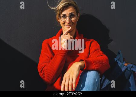 Frau mit rotem Pullover sitzt an einer schwarzen Wand, hält ihre Hand am Kinn und sieht erstaunt aus. Bild des Universitätskonzepts. Stockfoto
