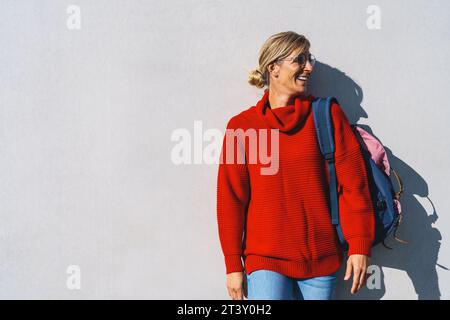 Porträt einer jungen Frau mit rotem Pullover und Rucksack, die nach rechts isoliert auf einer Betonwand mit Kopierraum schaut. Wunderschönes Studentenmädchen-Klo Stockfoto