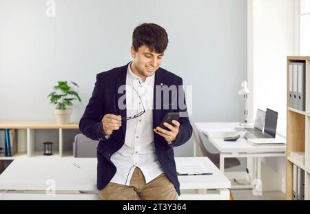 Zufriedener junger Geschäftsmann, der Handy per Telefon in der Nähe des Arbeitsplatzes in seinem Büro benutzt. Stockfoto