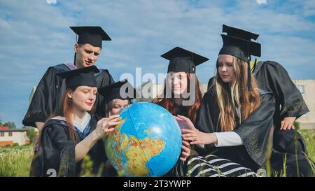 Absolventen in schwarzen Gewändern untersuchen einen geografischen Globus, der auf dem Gras sitzt. Stockfoto