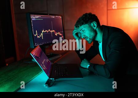 Nachtporträt eines seriösen, gutaussehenden Händlers, der ein Mobiltelefon hält, mit einem Laptop online arbeitet und Krypto-Charts im modernen Büro sitzt. Erfolgreich Stockfoto
