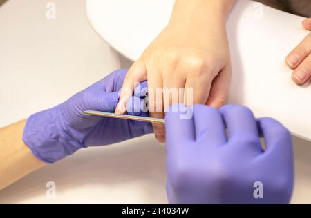 Maniküre Formen, feilen die Nägel einer schönen Frau, Finger halten. Ein weißer Manikürmeister arbeitet in Handschuhen an einem weißen Arbeitsplatz im Wellnesssalon. Weich Stockfoto