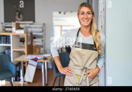 Fröhliche blonde Frau in grauem Oberteil und beigefarbener Schürze steht in einem Schneiderarbeitsplatz. Sie hat ein Maßband und eine Schere in ihrer Schürzentasche und ist s Stockfoto