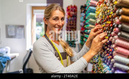 Fröhliche Schneiderin mit Maßband um den Hals wählt aus einer farbenfrohen Wand-Fadenanzeige Fäden für eine Nähmaschine aus Stockfoto