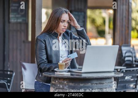 Junge frustrierte Buchmacherin, die im Straßencafé sitzt und den Kopf berührt, nachdem sie verloren hat, mit Plastikkarte zu wetten und auf den Bildschirm des Netbooks zu schauen Stockfoto