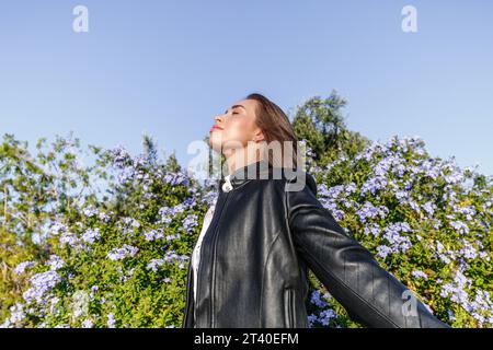 Hübsche junge Frau mit blonden Haaren und in einer Lederjacke gekleidet, mit geschlossenen Augen, die frische Luft im Wald an einem sonnigen Tag atmen Stockfoto