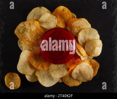 Blick von oben auf einen Haufen Kartoffelchips mit roter Dippsoße auf einem Servierbrett aus schwarzem Schiefer auf einem grauen Gipstisch. Auswahl Von Handwerklichen Chips Stockfoto