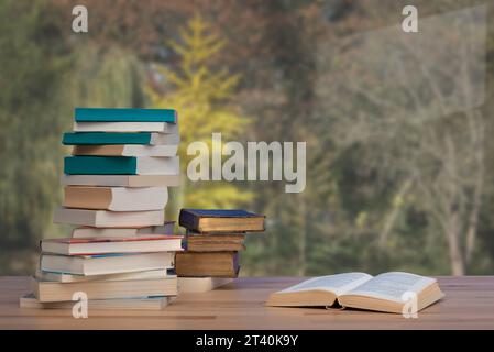 Stapel alter Bücher und geöffneter Roman auf Holztisch vor dem Fenster mit Herbstblick Stockfoto