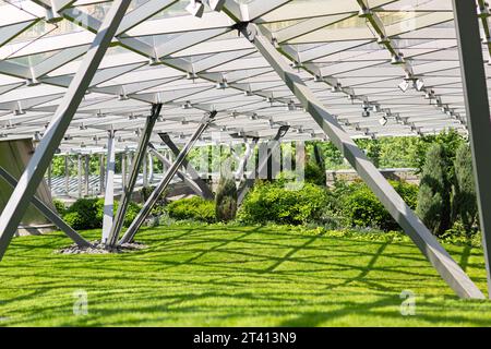 Ein Gewächshaus mit grünem Rasen und Büschen unter einer Kuppel aus Eisenpfählen und einem Glasdach, der Bau eines Wintergartens an einem sonnigen Tag. Stockfoto