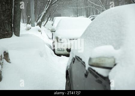 Eine Reihe geparkter Autos auf einer Stadtstraße an einem kalten Wintertag in der Nähe der Straße zwischen den Bäumen bedeckt ein Schneesturm die Fahrzeuge. Stockfoto