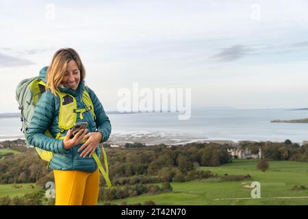 Eine junge Frau steht draußen mit einem Lächeln im Gesicht und einem Handy in der Hand Stockfoto