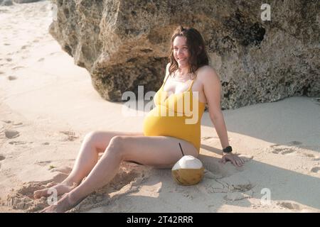 Eine glückliche schwangere Frau mit großem Bauch sitzt am Strand in der Nähe des Ozeans. Stockfoto