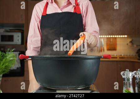 Eine Frau, die frische Karotte auf Hühnersuppe, Hühnerbrühe, in einem Topf hinzufügt. Gesundes Lebensmittelkonzept Stockfoto