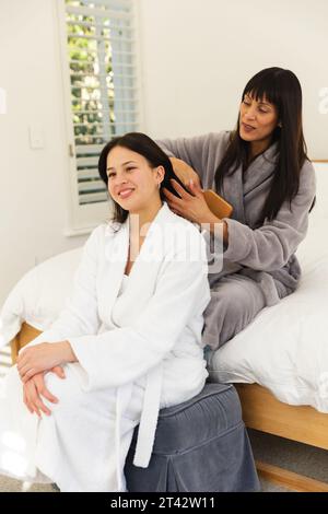 Glückliche, vielfältige Mutter und Tochter in Bademänteln, sitzen auf dem Bett und putzen zu Hause Haare, Kopierraum Stockfoto