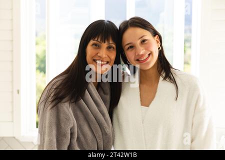 Glückliche, vielfältige Mutter und Tochter, die Bademäntel tragen und sich im Badezimmer zu Hause, Kopierraum umarmen Stockfoto
