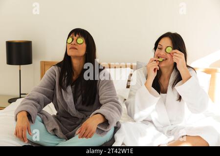 Diverse Mutter und Tochter in Bademänteln, sitzen auf Bett mit Gurken auf Gesichtern zu Hause, Kopierraum Stockfoto