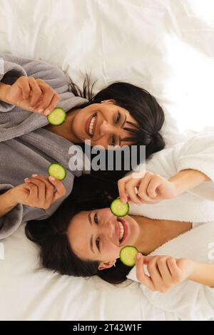 Diverse Mutter und Tochter in Bademänteln, liegend auf Bett mit Gurkenscheiben zu Hause, Kopierraum Stockfoto