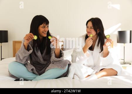 Vielfältige Mutter und Tochter in Bademänteln, auf dem Bett sitzen und Gurken essen zu Hause, Kopierraum Stockfoto