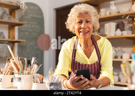 Glückliche zweirassige ältere Töpferin mit grauem Haar, mit Smartphone und lächelnd im Töpferstudio Stockfoto