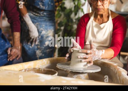 Glückliche zweirassige ältere Töpferin mit grauem Haar, mit Töpferrad im Töpferstudio Stockfoto