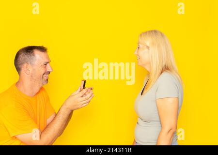 Mann, der einer Frau einen Antrag macht, die glücklich und überrascht aussieht. Das Konzept von Liebe und Ehe. Isolierter gelber Hintergrund. Romantisches Konzept, Schmuck kaufen Stockfoto