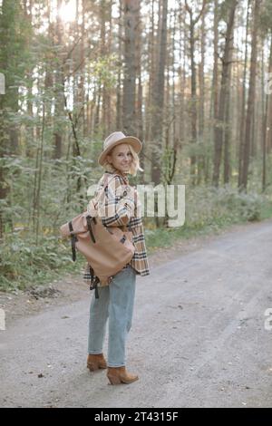 Lächelnde, stilvolle Frau, die im Wald steht und einen Rucksack trägt Stockfoto