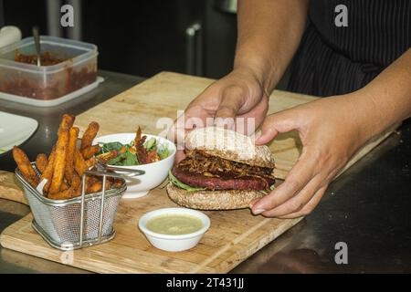 Nahaufnahme eines Küchenchefs, der einen Rote-Bete-Bohnen-Burger mit Süßkartoffelfritten, Salat und Dippsoße zubereitet Stockfoto