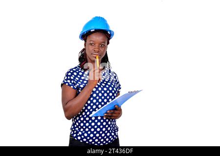 Eine junge Bauingenieurin trägt einen blauen Schutzhelm und denkt darüber nach, was sie auf die Zwischenablage schreiben soll. Stockfoto