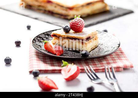 Ein Stück Napoleonkuchen mit Mohnsamen und Kirschen, dekoriert mit Erdbeeren und Blaubeeren auf hellem Hintergrund. Schließen Stockfoto