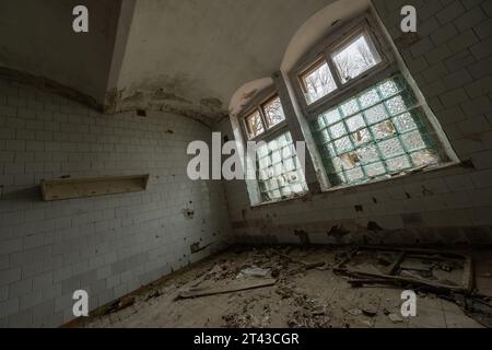 Niedriger Winkel des Innenraums der verlassenen geräumigen Halle mit Steinmauern, bogenförmigen Glasfenstern und verstreuten Müll-Möbeln Stockfoto