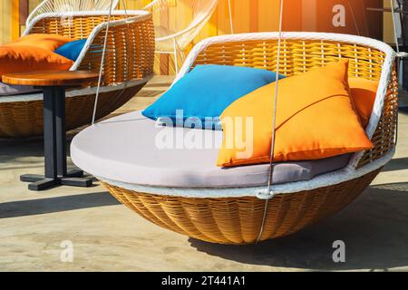 Farbenfrohe Kissen auf einem modernen Tagesbett. Braune Rattan-Liegestühle mit orangefarbenen und blauen Kissen, die mit Seilen auf der entspannenden Terrasse hängen. Sonnenliege zum Sonnenbaden und Stockfoto