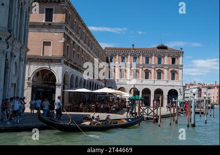 Eine Gondel vorbei an einem kleinen Platz mit einem Café im Freien und in der Nähe des Rialto Frischobst- und Gemüsemarktes am Ufer des Canal Grande in Venedig im Stockfoto