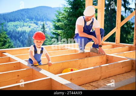 Vater mit Kleinkind-Sohn, der ein Holzrahmenhaus baut. Mann, der seinen Sohn in der Messung der Distanz mit Bandmaß auf der Baustelle anweist, mit Helmen und blauen Overalls an sonnigen Tagen. Stockfoto