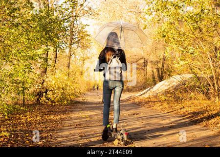 Ein Mädchen auf einem Elektroroller im Herbst unter einem transparenten Schirm fährt durch den Herbstwald entlang der Straße, spaziert im Herbst durch den Park, Stockfoto