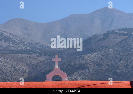 Bellfry und Kreuz auf einer kleinen Kirche zwischen Sissi und Malia mit Bergen im Hintergrund, Kreta, Griechenland, Europa. Stockfoto