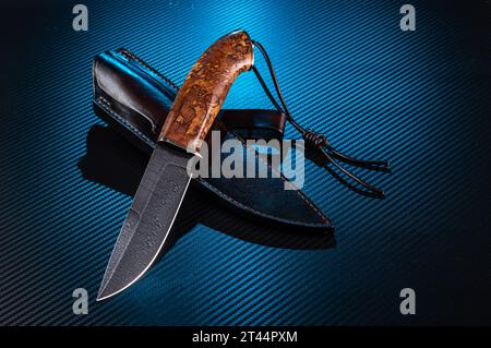 Jagdmesser aus Damaszener Stahl. Hochwertiges Messer mit Ledermantel. Messer und Lederschlaufe. Kohlehintergrund mit blauer Hintergrundbeleuchtung. Stockfoto