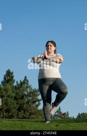 Übergewichtige Frau mittleren Alters übt Yoga im Freien barfuß, macht Gleichgewichtsübung auf einem Bein, Baumhaltung, blauer Himmel Hintergrund. Fitness, Yoga, Gewicht Stockfoto