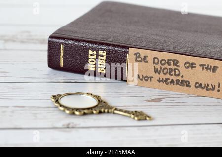 Sei Täter des Wortes, nicht nur Hörer, handgeschriebener Vers mit geschlossenem heiligem Bibelbuch und antikem goldenem Spiegel auf Holz. Eine Nahaufnahme. Jakobus 1:22 Schrift Stockfoto