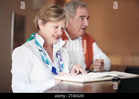 Seniorenpaar, liest und unterzeichnet Dokumente an der Hotelrezeption für Buchung, Reservierung oder Urlaub. Reifer Mann und Frau, die Formulare ausfüllen Stockfoto