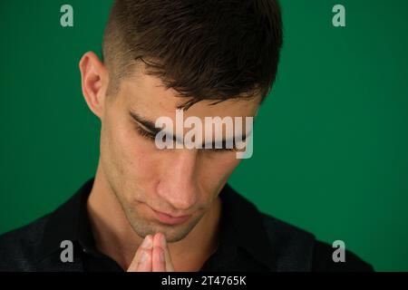 Emotionen eines gutaussehenden Mannes auf grünem Hintergrund, Chromakey-Nahaufnahme, dunkles Haar, junger Mann. Der Mann beugte sich vor Gott und betete zu Hause auf schwarzem Hintergrund. Hände im Gebet halten, Augen geschlossen. Stockfoto