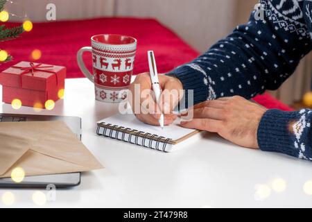 Schreiben Sie eine Weihnachtskarte. Männlicher Erwachsener macht Grußkarten im Heimbüro während Weihnachten. Mann Hände in blauem Pullover, der leeres Papier hält und umhüllt Stockfoto