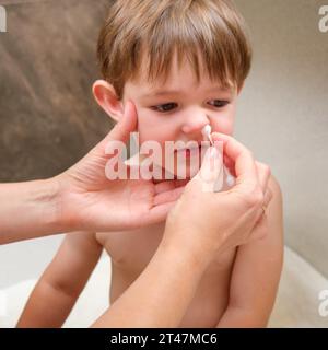 Die Mutter entfernt Rotz aus der Nase ihres kleinen Jungen mit einem Wattestäbchen im Badezimmer. Kind im Alter von zwei Jahren (zweijähriger Junge) Stockfoto