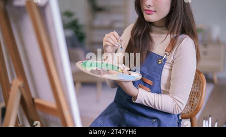 Junge asiatische Künstlerin mit Pinsel und Farbpalette, um Bilder auf Leinwand zu erstellen. Stockfoto