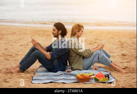 Lächelnder Junger Mann Und Frau Nachrichten Auf Smartphones Beim Picknick Im Freien Stockfoto