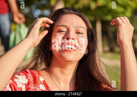 Frau, die ihr Sonnencreme ins Gesicht legt. Lächelnde junge Latina mit langen Haaren und Sonnencreme im Gesicht. Stockfoto