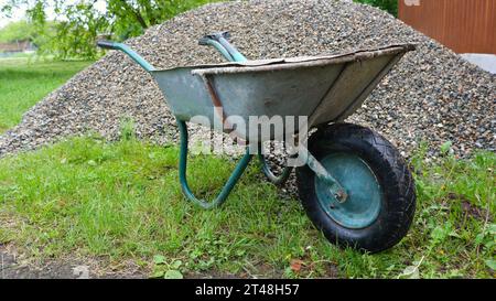 Alter Gartenwagen, der neben einem Haufen grauen Kies auf einem grünen Rasen in einer ländlichen Gegend steht, manueller Transport von zerdrücktem Stein auf einer Schubkarre Stockfoto