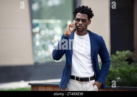 Selbstbewusster afroamerikanischer Geschäftsmann in elegantem Anzug und lässigem T-Shirt, der auf dem Handy spricht und in der Stadt spaziert. Trendige Darstellung moderner ci Stockfoto